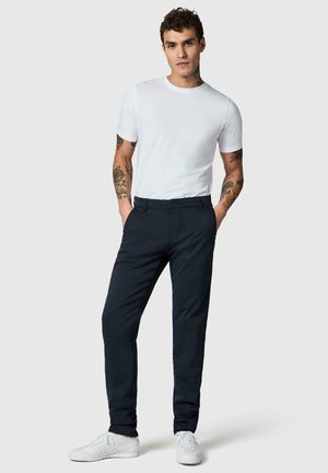 Giovane uomo con capelli ricci e tatuaggi che indossa una semplice maglietta bianca, pantaloni scuri aderenti e sneakers bianche, in piedi con le mani nelle tasche.