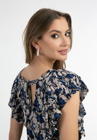 Blau-beiges gemustertes Kleid mit Rüschensäumen und einem Schlüsselauflösendetail auf der Rückseite. Modell trägt Perlenohrringe, heller Hintergrund.