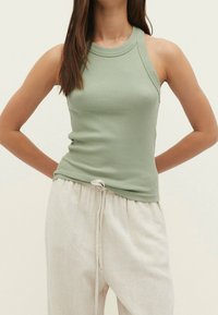 Vihreä urheilullinen toppi korkealla kauluksella, kapea malli ja leveät olkaimet, yhdistettynä beigekiristeisiin housuihin.