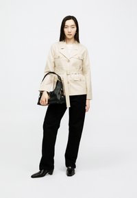 Γυναίκα που φοράει μπεζ σακάκι με ζώνη, μαύρο παντελόνι και μαύρες μπότες, κρατώντας μια μαύρη δερμάτινη τσάντα με χρυσές λεπτομέρειες.