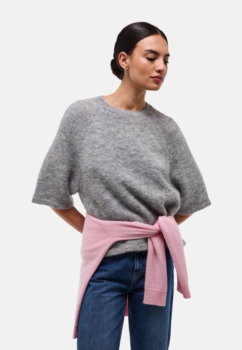 Femme aux cheveux foncés attachés, portant un pull gris à manches courtes, un jean bleu et un pull rose noué à la taille.