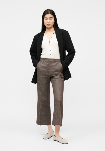 Femme debout les mains dans les poches, portant un blazer noir, un haut blanc boutonné, un pantalon marron raccourci et des chaussures beige sans lacets.
