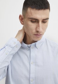 Camisa de rayas azul claro y blanca con cuello abotonado, mangas largas y puños con botón. La tela parece suave con una textura lisa.