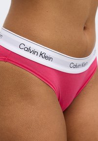 Ružové bavlnené spodné prádlo s bielym pásom obsahujúcim čierne logo „Calvin Klein“. Hladká textúra a jednoduchý dizajn bez ďalších vzorov.