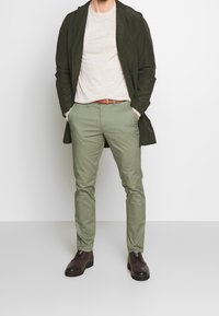 Olivgrön lång kappa, ljusbeige t-shirt och ljusgröna byxor; bruna läderskor med snördetaljer, brun bälte, skräddarsydd passform.