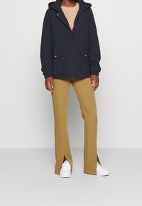 Námořnická bunda s kapucí, dvěma předními kapsami a kontrastním červeným zipem. Béžové kalhoty s postranními rozparky, doplněné bílými teniskami.