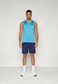 Ανοιχτό μπλε αμάνικο αθλητικό μπλουζάκι, συνδυασμένο με ναυτικά σορτς. Λευκά αθλητικά παπούτσια και αθλητικές κάλτσες ολοκληρώνουν το ντύσιμο. Ο λογότυπος της Under Armour είναι ορατός.