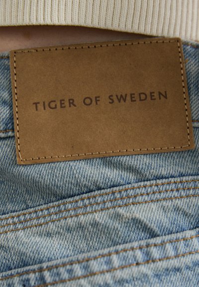Zil poga gaisz ziliem audumiem ar redzamiem diegiem; taisnstūra brūnas ādas uzlīme ar uzrakstu "TIGER OF SWEDEN" uz jostas.