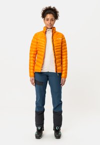 Feloranje donsjack met horizontale quilting, gecombineerd met blauwe broek met zwarte accenten en skischoenen. Soepele, lichte materialen.