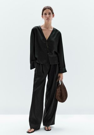 Zwarte satijnen blouse met lange mouwen en een diepe V-hals, afgewerkt met knoopdetails, gecombineerd met losse zwarte broeken. Het model houdt een gestructureerde bruine tas vast.