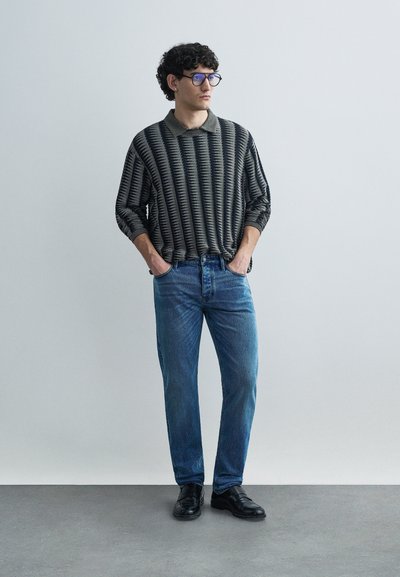 Πλεκτό πόλο πουκάμισο με μαύρες και γκρι ρίγες, σε χαλαρή εφαρμογή, συνδυασμένο με ανοιχτό μπλε τζιν με ίσια γραμμή και μαύρα δερμάτινα παπούτσια.