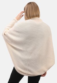 Une personne avec les cheveux blonds courts portant un poncho en tricot côtelé crème sur un pantalon noir, tournant le dos et touchant sa tête d'une main.