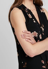 Žena se zkříženýma rukama, oblečená v bezramínkovém černém topu s béžovým a hnědým květinovým vzorem na jednobarevném pozadí.