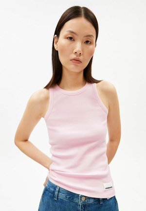 Jeune femme aux cheveux bruns lisses portant un haut côtelé sans manches rose clair et un jean bleu sur un fond uni.