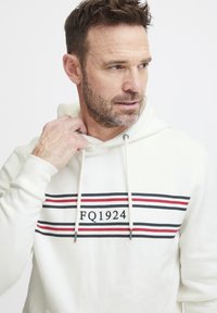 Sudadera blanca con un logo frontal "FQ1924" y rayas horizontales en rojo y negro. Presenta cordones y una textura suave.