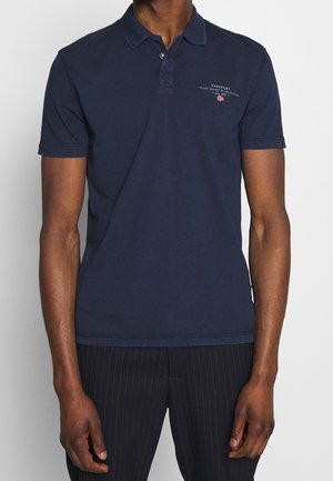 Man wearing navy blue short-sleeve polo shirt with small logo and text on left chest, paired with black pinstripe trousers.