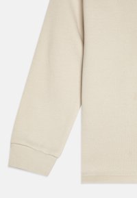Indumento a maniche lunghe color beige chiaro con polsini e orlo a coste. Tessuto morbido e testurizzato con un design semplice e lineare.