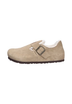 Zapato slip-on de ante beige con un suave forro de piel de pelo y una correa ajustable. Presenta una suela texturizada y un sutil logotipo en el lateral.