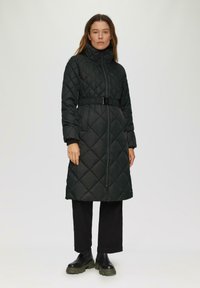Zwarte, gewatteerde lange jas met hoge kraag, riem in de taille en ritssluiting. Bevat een ruitpatroon en geribde mouwen, gedragen over zwarte broek.