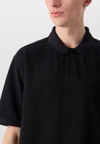 Sort polo-shirt lavet af bomuld, med en knappet krave og korte ærmer. Tekstureret stof med et broderet logo på brystet.