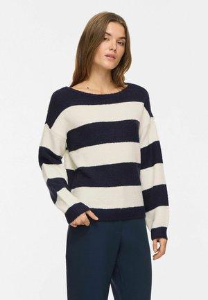 Ung kvinde iført en marineblå og hvid bredstribet sweater og mørke bukser, stående mod en ensartet lys baggrund.