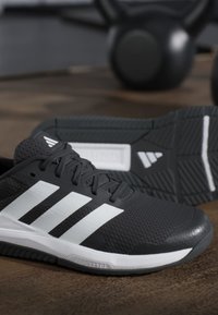 Chaussures de sport noires et blanches avec trois bandes blanches sur le côté, placées sur un sol en bois avec des kettlebells en arrière-plan.