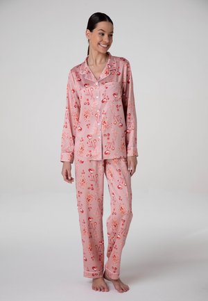 Donna sorridente che indossa un completo pigiama rosa floreale a maniche lunghe con camicia abbottonata e pantaloni coordinati, in piedi a piedi nudi su sfondo uniforme.
