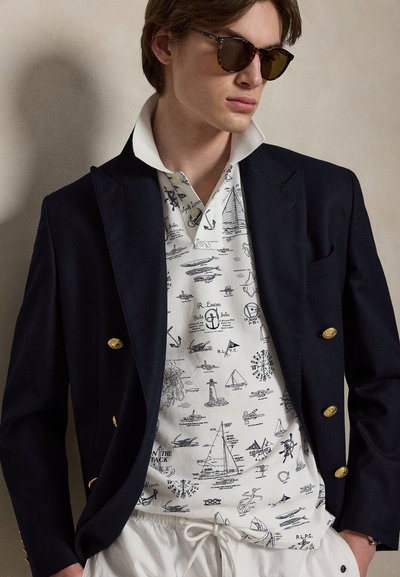 Νέος άνδρας που φοράει σκούρο σακάκι με χρυσά κουμπιά πάνω από ένα λευκό πουκάμισο με ναυτικό σχέδιο, λευκό παντελόνι με κορδόνι και γυαλιά ηλίου από χελώνα.