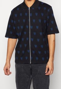 Maglietta nera a maniche corte con zip, caratterizzata da un motivo a cuori nei toni del blu e del verde. Collo a polo e vestibilità comoda.