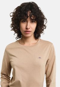 Beige Langarm-T-Shirt aus weichem Stoff, mit Rundhalsausschnitt und kleinem gesticktem Logo auf der linken Brust.