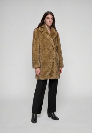 Kvinde iført en brun faux fur frakke, sorte bukser med lige ben og sorte ankelstøvler med hæl, stående mod en ensfarvet lys baggrund.