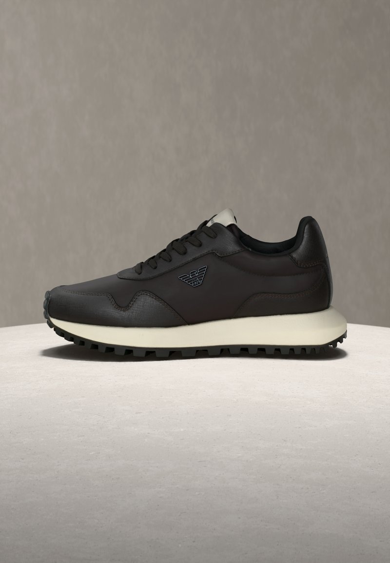 Temno rjav usnjen športni copat z reliefnimi poudarki, črnimi vezalkami, kremasto barvo podplata in logotipom blagovne znamke na strani, prikazan na svetli površini.