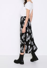 Nainen, jolla on päällään valkoinen lyhythihainen paita, musta maxi-hame, jossa on valkoinen lehtikuvio ja halkio, sekä mustat paksupohjaiset nilkkasaappaat, seisoo sivuttain.