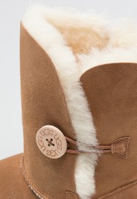 Botte UGG en daim marron avec doublure en laine blanche et un bouton rond cousu portant l'étiquette « UGG » fixant le côté.