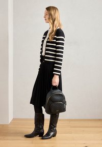 Cardigan rayé noir et blanc, jupe plissée noire, bottes de cowboy noires et un sac à dos texturé noir avec des accents dorés.
