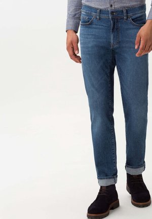 Homme portant un jean bleu retroussé, une chemise grise à carreaux et des bottes noires en daim à lacets, debout sur un fond clair.