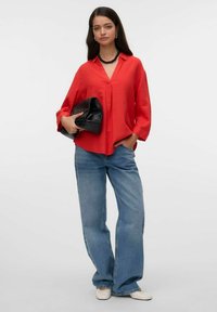 Blouse rouge à manches longues avec un décolleté en V et des plis, associée à un jean large bleu clair. Le mannequin tient une pochette noire.