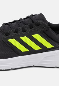 Chaussure de sport noire avec une finition texturée, arborant trois bandes jaunes audacieuses, un design à lacets et une semelle intermédiaire blanche.