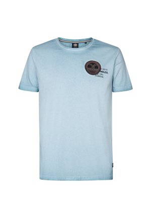 Helles blaues T-Shirt aus Baumwolle mit rundem Ausschnitt und kurzen Ärmeln. Ein runder brauner Patch ist an der oberen linken Seite aufgestickt.
