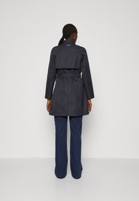 Impermeabile blu navy con cintura, collo strutturato e sovrapposizione sulle spalle, dotato di maniche lunghe e una finitura liscia e resistente all'acqua.