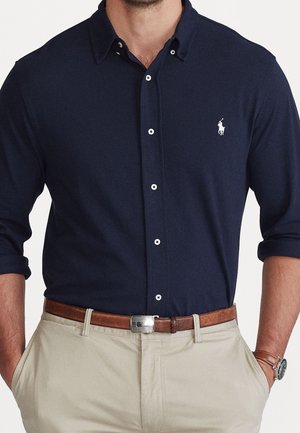 Mężczyzna w granatowej koszuli zapinanej na guziki z białym logo, beżowych spodniach, brązowym skórzanym pasku i zegarku na ręku z paskiem ze skóry, ręce w kieszeniach.