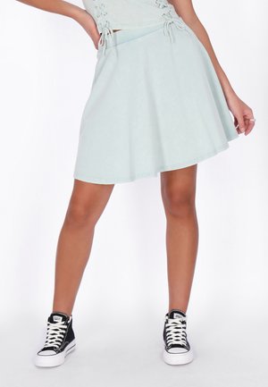 Femeie purtând o fustă bleu deschis și un top asortat, cu adidași negri înalți, stând cu o mână pe șold.