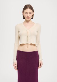 Cardigan scurt din tricot cu nervuri, de culoare crem, cu mâneci lungi și decolteu în V. Are un fermoar frontal și un tiv ondulat. Asortat cu o fustă purpurie.