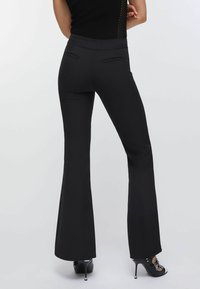 Pantalon noir évasé avec une texture lisse, une coupe ajustée et des poches arrière. Porté avec des talons ouverts noirs.