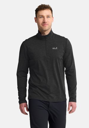 Man draagt een donkergrijze lange mouwen trui met rits en een klein logo op de borst en zwarte broek tegen een effen achtergrond.