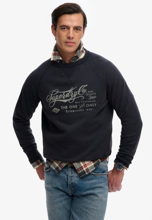Mann trägt dunkelblauen Pullover mit dem Text "Superdry Co.", darunter kariertes Hemd, blaue Jeans und Armbanduhr, steht mit den Händen in der Hüfte.