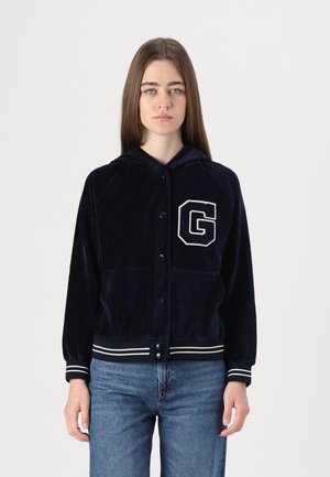 Marineblauwe velours jacket met capuchon, voorzien van een witte letter "G" embleem, drukknoopsluiting, twee voorzakken en gestreepte manchetten en zoom.
