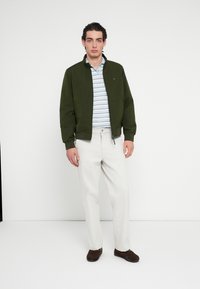 Jachetă bomber verde peste un tricou polo cu dungi albastre și albe, asortată cu pantaloni bej deschis și pantofi maro. Design simplu și modern.