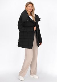Sort puffjakke med høj krave, med en quiltet design og lynlåslukning. Båret over en hvid top og beige bukser.