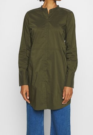 Personne portant une tunique manches longues vert olive avec un col officier et un jean bleu sur un fond uni.